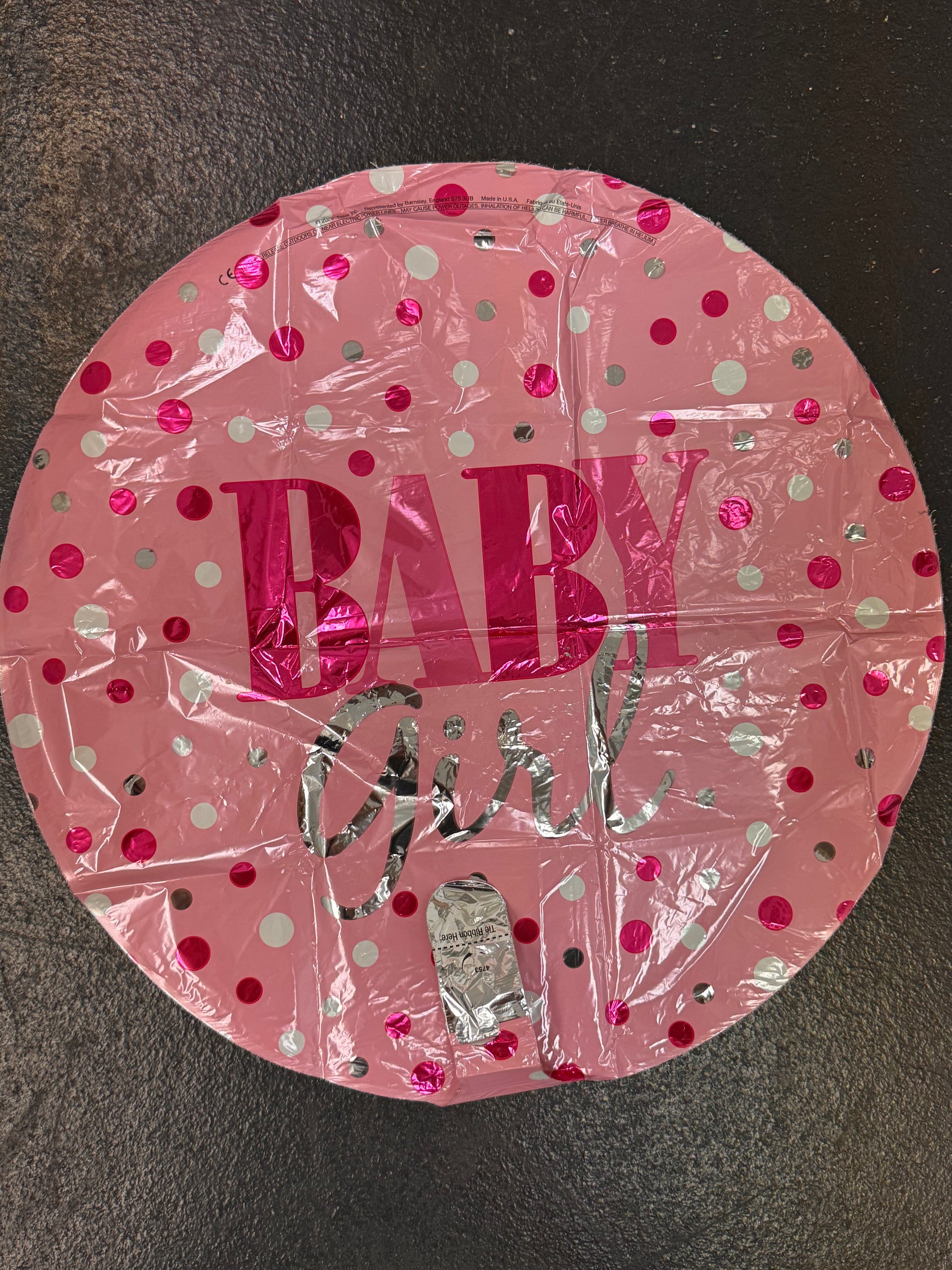 A round pink foil balloon with 'BABY girl' printed on it, decorated with shiny pink, silver, and white polka dots, shown on a dark textured surface.
