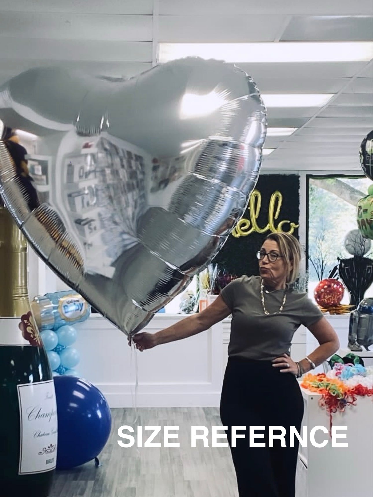 Woman holding a large silver heart-shaped balloon in an indoor setting with 'SIZE REFERENCE' text overlay.
