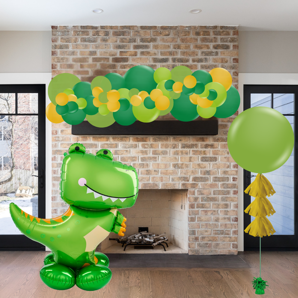 Green dinosaur balloon and green balloons with yellow tassels in front of a brick fireplace.