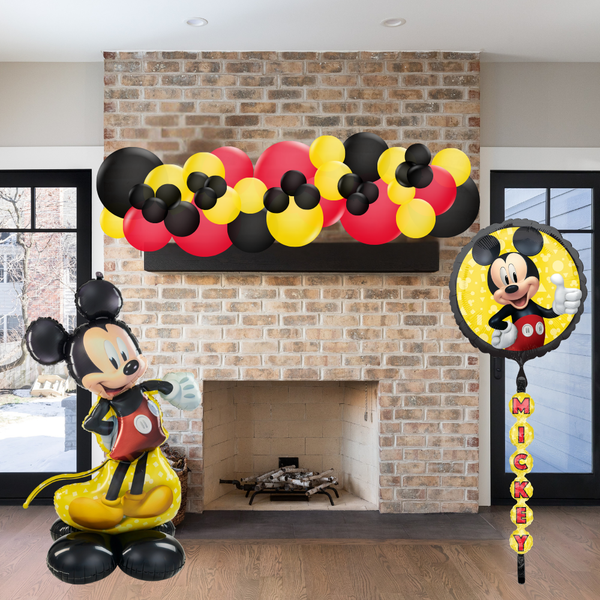 Mickey Mouse-themed balloons and decorations in front of a brick fireplace.